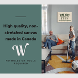 Lifestyle image of the horizontal, 20x30” inch easy to hang canvas wall art hung in a living room above a couch with a man sitting on his phone while his son vacuums the carpet around them with graphic saying "High quality, non-stretched canvas made in Canada"